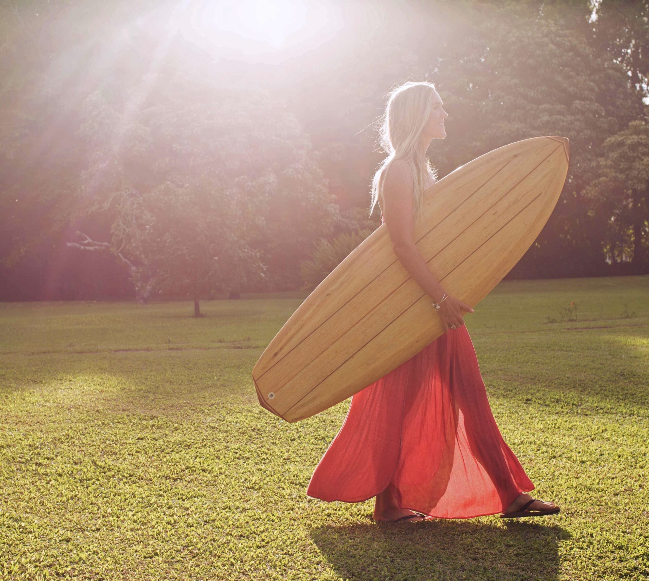 Bethany walks through the grass with a wooden surfboard under her arm.